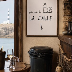 Peut inclure: Une affiche encadrée avec le texte "ça va à LA JAILLE" et une illustration d'une bouteille et d'un gobelet avec une paille. Une poubelle noire est au premier plan. Un phare est visible à travers une fenêtre.