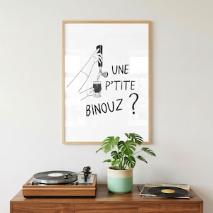 Peut inclure: Une affiche encadrée en noir et blanc avec le texte "UNE P'TITE BINOUZ ?" représente une main versant une boisson dans un verre. L'affiche est dans un cadre en bois. Une platine et une plante en pot sont au premier plan.