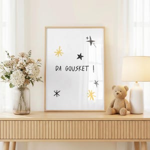 Peut inclure: Une affiche encadrée avec le texte "DA GOUSKET!" et des illustrations d'étoiles noires et dorées. L'affiche est exposée sur une console en bois, à côté d'un ours en peluche, d'une lampe et d'un vase de fleurs.