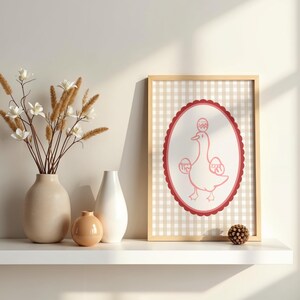 Peut inclure: Une affiche encadrée avec un dessin d'un canard rose tenant des œufs. L'œuvre est placée sur un fond à carreaux beige et blanc, encadrée de bois clair. Des vases et des fleurs sont sur une étagère blanche.
