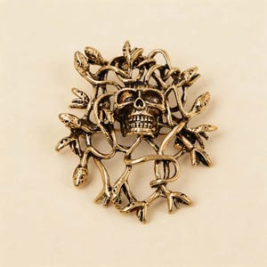 May include: A gold-toned metal brooch featuring a central human skull surrounded by a detailed arrangement of intertwined snakes. The design has an antique finish with dark recessed areas that highlight the texture of the skull and snake scales.