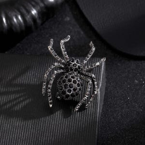 Op de afbeelding: Een spinvormige broche gemaakt van donkergrijs metaal, met een rond achterlijf en borststuk ingelegd met talloze kleine, gefacetteerde zwarte kristallen. De acht gelede poten zijn eveneens gedetailleerd met rijen zwarte kristallen.
