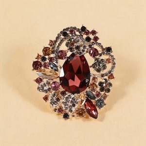 Pode incluir: Um broche decorativo com um grande cristal central em forma de gota de cor vermelho-escura, rodeado por cristais mais pequenos redondos, marquise e em forma de folha em tons de bordeaux, âmbar, cinzento e preto, numa armação de metal dourado.