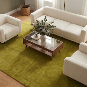 Puede incluir: Un conjunto de sala de estar con un sofá de bouclé color crema y dos sillones a juego dispuestos alrededor de una mesa de centro rectangular de madera con tapa de cristal. Una gran alfombra verde oliva se sitúa bajo los muebles sobre el suelo.