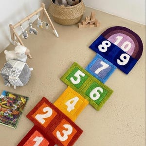 May include: A colorful hopscotch rug featuring a rainbow-inspired design with white numbers one through ten tufted onto square and arched segments. The rug displays a gradient of red, orange, yellow, green, blue, and purple sections.