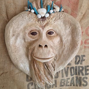 Puede incluir: Una cara de orangután esculpida con barba y una corona de conchas marinas y detalles turquesas. La cara es de color beige claro con ojos marrones oscuros y una expresión sonriente. El fondo es un saco de arpillera.