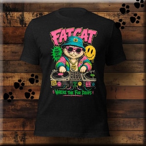 May include: A black t-shirt featuring a graphic of a cat wearing a blue bucket hat, pink sunglasses, and a gold chain. The cat is DJing on a turntable console. Text includes FAT CAT, DROP PAWS NOT BOMBS, PAW BEATS, RAVE MEOW, ACID 4 LIFE, BASS IS PURR, and WHERE THE FUR DROPS.