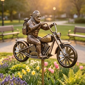 May include: Bronze-colored garden stake featuring a Bigfoot figure riding a motorcycle. The motorcycle has the words "Yeti Riders" on the gas tank. The Bigfoot is wearing sunglasses and a headband. The stake is in a garden with flowers and benches.