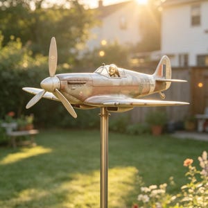 May include: A detailed metal airplane weathervane with a propeller and pilot figure. The weathervane is mounted on a metal pole in a grassy yard, with a blurred background of trees and a house. The plane has a weathered, vintage appearance.