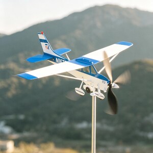 May include: A blue and white model airplane with a spinning propeller. The plane is mounted on a metal rod, with the number "172" visible on the side. The tail has a red accent.