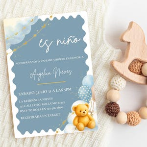 May include: A rectangular baby shower invitation card with a scalloped edge, featuring a dusty blue background, a golden moon and star garland, a brown teddy bear, and blue balloons. The text reads: es niño, ACOMPAÑANOS A UN BABY SHOWER EN HONOR A Angelica Nieves, SABADO, JULIO 31 A LAS 5PM, LA RESIDENCIA NIEVES, 123 CALLE OSO, ROLLA MO 12345, RSVP TO ROSA EN 123-456-7890, REGISTRADA EN TARGET.