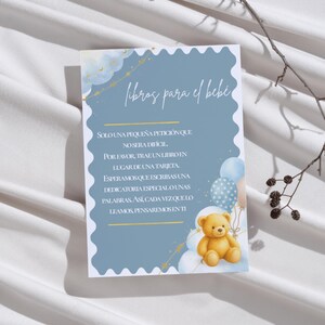 May include: A rectangular card with a scalloped white border on a dusty blue background. The text reads "libros para el bebé" followed by a request to bring a book instead of a card. The design features a golden teddy bear, blue balloons, and stars.