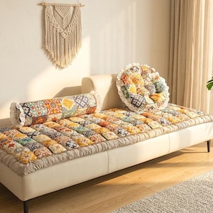 Op de afbeelding: Een crèmekleurige daybed met een kleurrijke patchwork quilt en bijpassende bolster en rond kussen. De kamer heeft een macramé wandkleed, een potplant en een kleine houten bijzettafel met vazen.