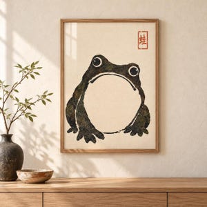 Op de afbeelding: Een ingelijste wandkunstprint met een donkergroene, getextureerde illustratie van een ronde kikker met grote ogen. De achtergrond is lichtcrème van kleur en in de rechterbovenhoek staat een kleine rode stempel met het Chinese karakter voor kikker.