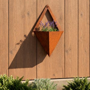 Può includere: Una fioriera da parete a forma di diamante realizzata in metallo arrugginito con una patina arancione-marrone usurata. Il contenitore geometrico contiene un mazzo di fiori di lavanda viola ed è fissato a una superficie in legno con una vite.