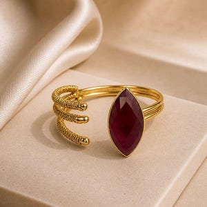 Op de afbeelding: Een goudkleurige open ring met een dieprode edelsteen in marquise-slijpvorm als middelpunt. De ringband bestaat uit drie parallelle draden, waarbij de uiteinden aan één kant een textuurrijk, spiraalvormig ontwerp hebben met kleine afgeronde punten.