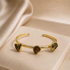 Könnte beinhalten: Ein goldfarbenes Armband in offener Form mit drei unregelmäßigen, dunkelbraunen, roh geschnittenen Steinen, die in polierten Metallfassungen sitzen. Das Armband besteht aus einem glatten, abgerundeten Reifen mit zwei kleinen Endkappen.