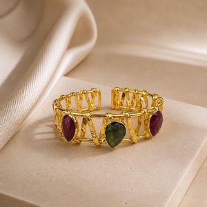 Könnte beinhalten: Ein goldfarbenes, offenes Armband mit einer käfigartigen Drahtstruktur und einer texturierten Oberfläche. Das Armband ist mit drei tropfenförmigen Edelsteinen verziert, darunter zwei dunkelrote Steine an den Seiten und ein dunkelgrüner Stein.
