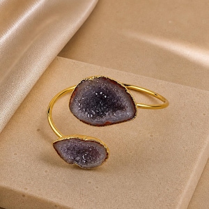 Könnte beinhalten: Ein goldfarbenes Armband in offener Form mit zwei asymmetrischen, rohkantigen violetten Geodenscheiben an den Enden. Die Geodensteine haben eine kristalline, texturierte Oberfläche in tiefen Violetttönen, während das Metallband glatt poliert ist.