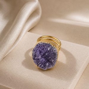 Könnte beinhalten: Ein goldener Ring mit einem großen, kreisförmigen, violetten Druzy-Edelstein als Mittelstück mit einer texturierten, kristallinen Oberfläche. Die Ringschiene besteht aus zwei dünnen, parallelen, goldfarbenen Metalldrähten.