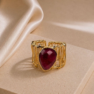 Könnte beinhalten: Ein goldfarbener Ring mit einem großen, facettierten, tief magentafarbenen tropfenförmigen Stein in der Mitte. Die Ringschiene besteht aus mehreren dünnen, vertikalen goldfarbenen Drähten, die eine offene, käfigartige Struktur bilden.