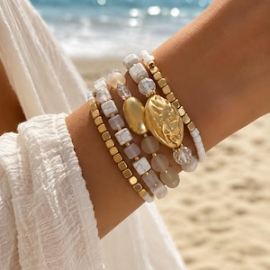 Könnte beinhalten: Ein Stapel aus fünf Perlenarmbändern mit einer Mischung aus weißen, marmorierten Perlen, durchscheinenden mattierten Perlen, kleinen goldfarbenen quadratischen Perlen, klaren facettierten Perlen und einem ovalen goldfarbenen Anhänger.