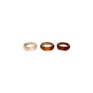 Puede incluir: Tres anillos dispuestos en una fila horizontal. El primer anillo es de color blanco roto translúcido con una superficie texturizada. El segundo anillo es de color ámbar intenso con una forma geométrica facetada. El tercer anillo es una banda lisa de color marrón oscuro.