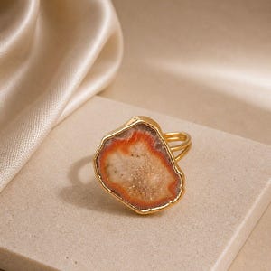 Könnte beinhalten: Ein goldener Ring mit einer großen, unregelmäßigen, rohen Scheibe aus Achatstein. Der Stein hat eine texturierte, kristalline Mitte in Beige- und Cremetönen, umgeben von einem leuchtenden, tief orange-roten Rand. Der Metallring ist dünn und goldfarben.