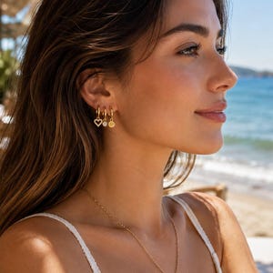 May include: Three gold hoop earrings worn in a single ear, each featuring a small dangling charm. The charms consist of a heart shape with embedded stones, a circular stone, and a plain gold disc. A thin gold chain necklace is also visible.