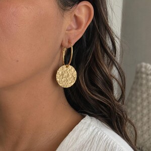 Op de afbeelding: Een gouden oorring met een ronde hanger met een getextureerde, gehamerde afwerking. De oorbel bestaat uit een dunne, gepolijste gouden ring die verbonden is met een platte, ronde schijf met een onregelmatig, reflecterend oppervlak.