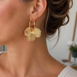 Peut inclure: Une boucle d'oreille créole dorée ornée d'une breloque fleur superposée et texturée avec des détails de pétales finement gravés. La finition dorée métallique reflète la lumière, soulignant le motif de surface aux lignes fines du design floral.