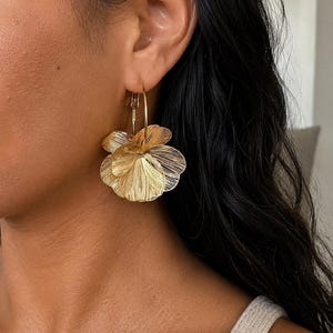 Peut inclure: Une boucle d'oreille créole dorée ornée d'une breloque florale en éventail à plusieurs niveaux. La breloque se compose de multiples pétales métalliques fins avec une texture de lignes gravées, suspendue à un anneau simple passant par le lobe.