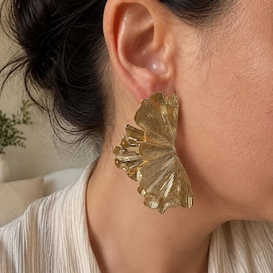 Peut inclure: Une grande boucle d'oreille dorée en forme d'éventail plissé avec une finition métallique texturée. Le bijou présente une forme semi-circulaire large qui s'étend depuis le lobe de l'oreille, mettant en valeur de fines lignes verticales.