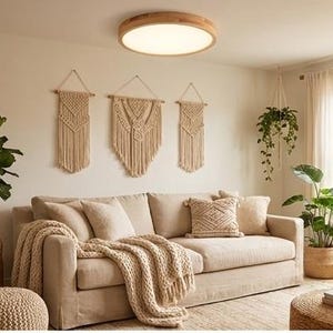 May include: A beige living room with a sofa, throw pillows, and a knitted throw. Three macrame wall hangings are above the sofa. A round wooden ceiling light and potted plants complete the cozy, neutral-toned space.