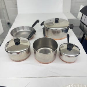 May include: A set of stainless steel cookware including a large lidded pot, a frying pan, and two smaller saucepans with lids. Each piece features a polished silver finish, black handles, and a distinct copper-colored base band for heat conduction.