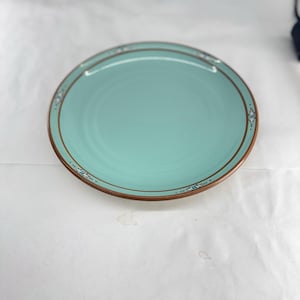 May include: A round, light turquoise dinner plate featuring a thin, dark brown rim and a decorative border pattern consisting of small, symmetrical blue and white geometric motifs spaced evenly around the edge of the plate's surface.