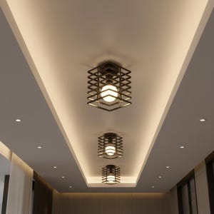 May include: Three modern ceiling lights with a geometric cage design. Each light fixture features a square, dark metal frame encasing a spherical, glowing light bulb. The lights are mounted on a white ceiling with recessed lighting.