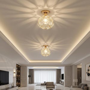 May include: Two gold-toned, crystal ceiling lights are mounted on a white ceiling. The lights cast a decorative pattern on the ceiling. The room has a modern design with marble flooring and a large television.