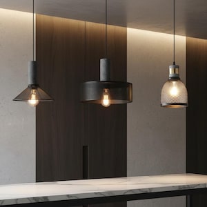 Könnte beinhalten: Drei moderne Pendelleuchten mit freiliegenden Glühbirnen. Die Lampenschirme bestehen aus Metallgitter und dunkelgrauen Materialien. Die Leuchten hängen von der Decke und beleuchten eine Marmorarbeitsplatte. Der Hintergrund zeigt dunkle Holzverkleidungen und eine hellfarbige Wand.