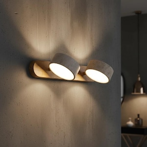 Può includere: Un moderno apparecchio a parete con due faretti orientabili. Le teste delle luci sono di colore grigio chiaro e montate su una base di legno. L'apparecchio è montato su una parete grigia strutturata, proiettando una luce calda.