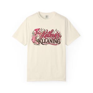 以下が含まれることがあります： ピンクとブラウンの文字で「Kathy's KLEANING」と書かれたグラフィックデザインが施された、クリーム色の半袖Tシャツ。デザインには、ギンガムチェックのリボンが付いたピンクのスプレーボトル、小さな花、泡、星のアクセントが含まれています。