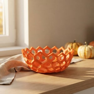 Puede incluir: Un cuenco de color naranja brillante con un patrón geométrico de panal de abeja y recortes hexagonales abiertos en toda la estructura. El cuenco tiene una base circular y un borde superior dentado, creando un aspecto ligero y enrejado.