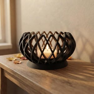 Pode incluir: Um suporte de vela preto mate com uma estrutura geométrica esférica de treliça aberta. O suporte possui uma base circular e contém um pequeno copo votivo de vidro transparente com uma vela de chá branca acesa no seu interior.