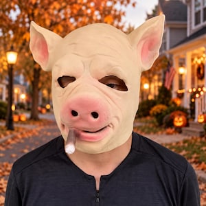 Könnte beinhalten: Eine Schweinemaske für den gesamten Kopf in hellem Rosaton mit großen spitzen Ohren und einer markanten Schnauze. Die Maske zeigt eine realistisch wirkende Zigarre, die aus dem Mundbereich ragt, sowie dunkle Augenöffnungen und Gesichtsfalten.