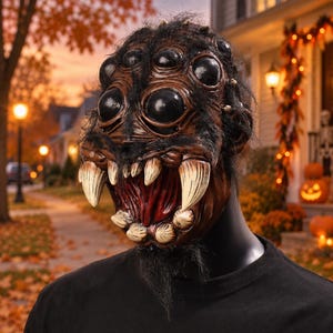 Op de afbeelding: Een bruin spinnenmasker met meerdere grote zwarte ogen, een getextureerde huid en opvallende gebroken witte hoektanden. Het masker is bedekt met donker, dun haar en heeft een wijd open mond die een dieprode binnenkant onthult.