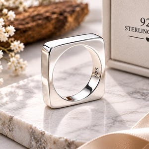 May include: A square-shaped sterling silver ring with a polished finish. The interior of the band is stamped with the text 925. The ring is presented near a box featuring the text 925 STERLING SILVER with a small heart icon between two lines.