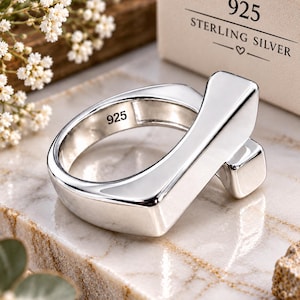 May include: A polished sterling silver ring with a thick, angular band design. The interior of the band is clearly stamped with the number 925. The ring is positioned near a beige box featuring the text 925 STERLING SILVER with a small heart icon.