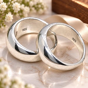 May include: Two polished silver rings with a smooth, rounded, thick band design. Each ring features the hallmark 925 stamped clearly on the inner surface, indicating sterling silver composition. The rings are positioned side by side.