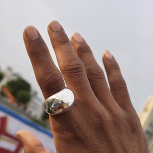 May include: A polished silver-tone ring with a wide, smooth, and rounded band. The surface of the ring is highly reflective, showing a clear mirror-like finish that captures the surrounding environment in its curved, metallic exterior surface.