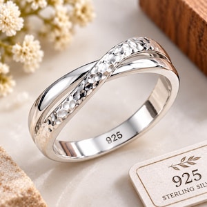 Puede incluir: Un anillo de plata con un diseño cruzado superpuesto, donde una banda presenta una textura martillada y la otra un acabado liso y pulido. El interior de la banda está claramente marcado con el número 925, indicando plata de ley.